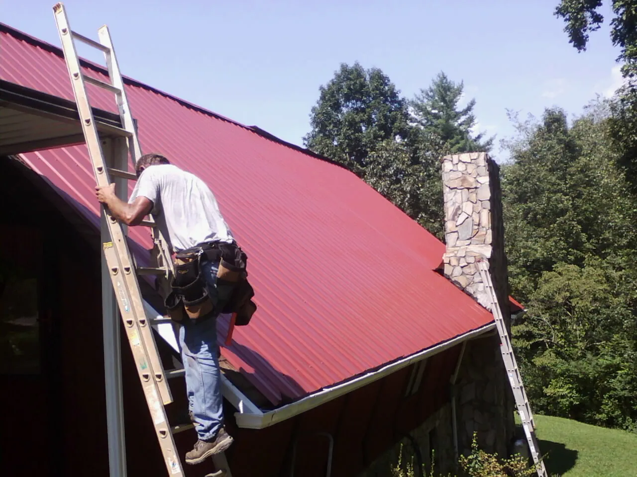 Roofer on a ladder beside a red metal roof and stone chimney — Soffit & Fascia Repair by Roofing Brothers in Meadville