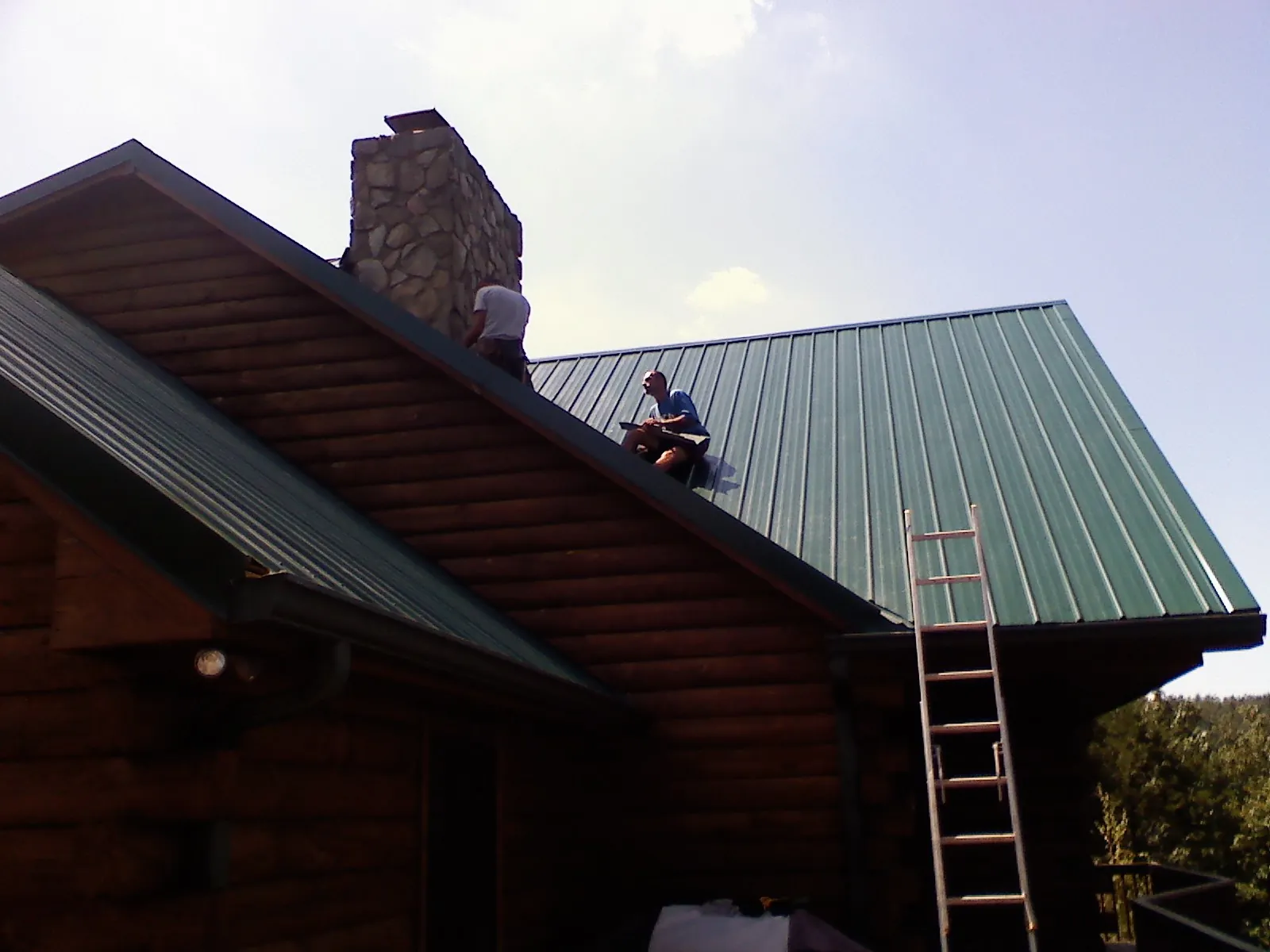 Roofers installing green metal panels beside a stone chimney on a log home in Meadville